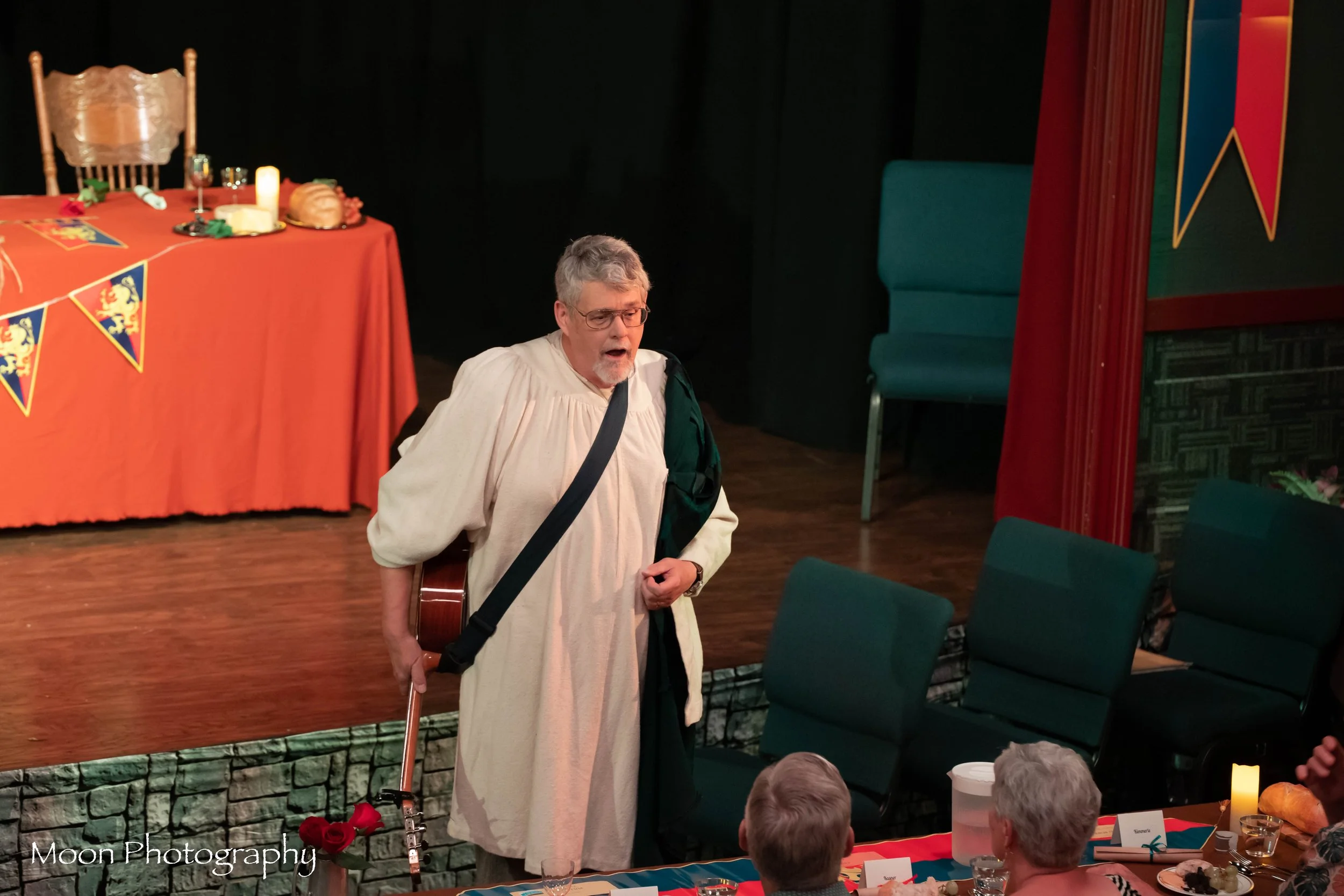 A man dressed as a priest with an acoustic guitar standing in front of a table with food and drinks at a celebration or event.