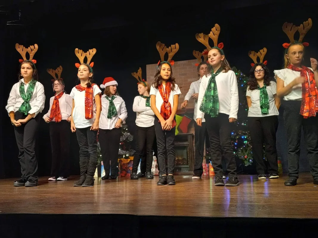 Group of children dressed in Christmas costumes with reindeer antlers and scarves, standing on stage during a holiday performance.