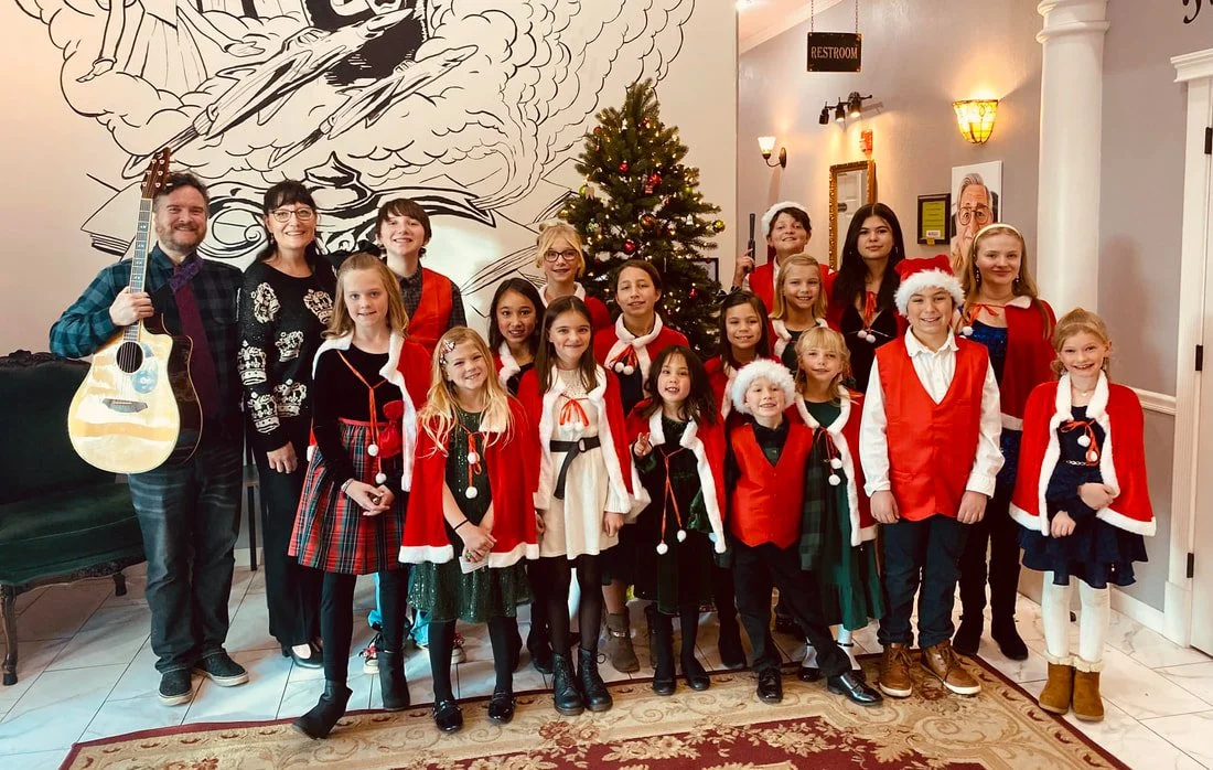 Group of children and adults celebrating Christmas indoors, with a decorated Christmas tree in the background. Many children are wearing Christmas costumes or red and green outfits, some with Santa hats. One adult on the left holds a guitar.