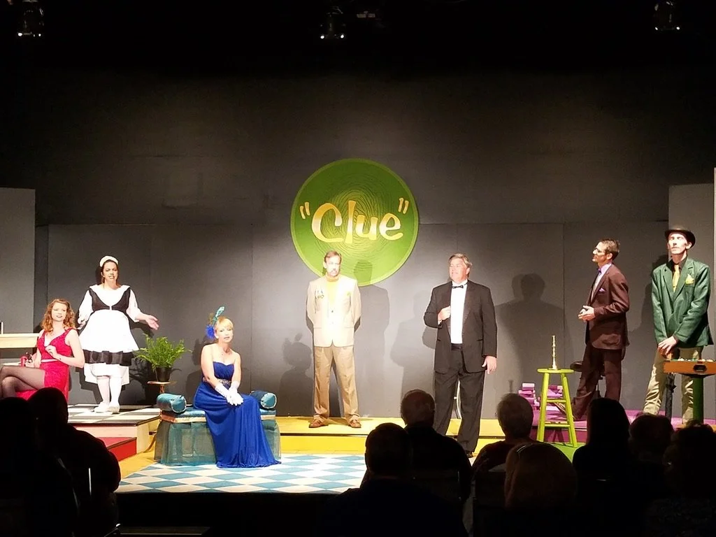 Stage scene from a production of the musical 'Clue' with actors dressed as characters including a maid, a woman in a blue dress, a man in a beige suit, a man in a tuxedo, and others, with a large 'Clue' sign in the background.