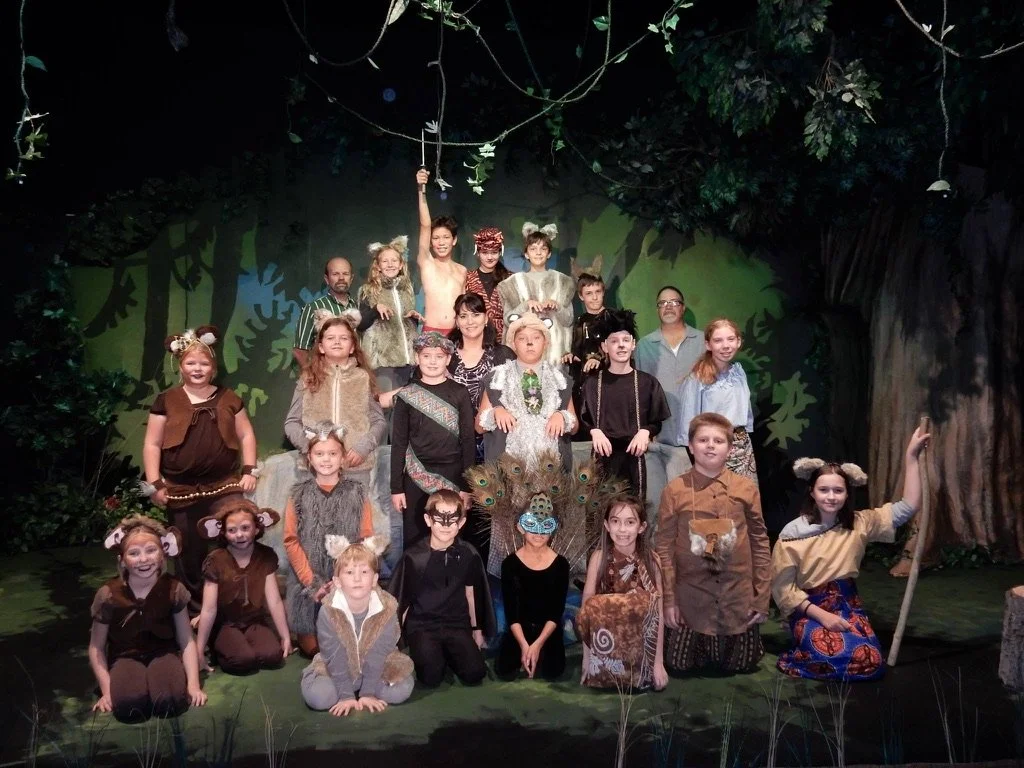 Children in costume on stage during a theatrical performance with jungle-themed backdrop and props.