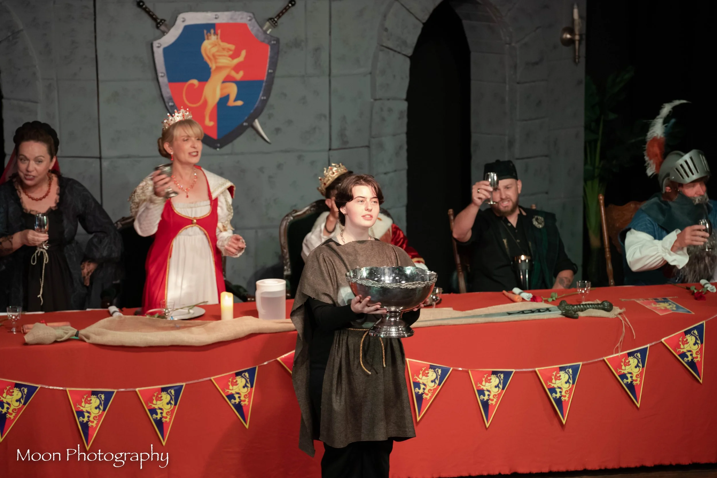 People dressed in medieval costumes sit at a long table decorated with banners featuring a lion and blue background, with a person in medieval attire holding a large silver bowl in front of them.