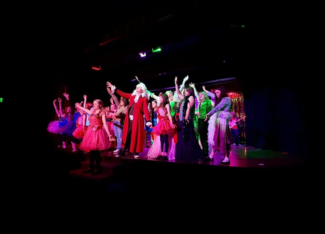 Children and adults performing a stage play or musical, dressed in colorful costumes, with a dark background and stage lights.