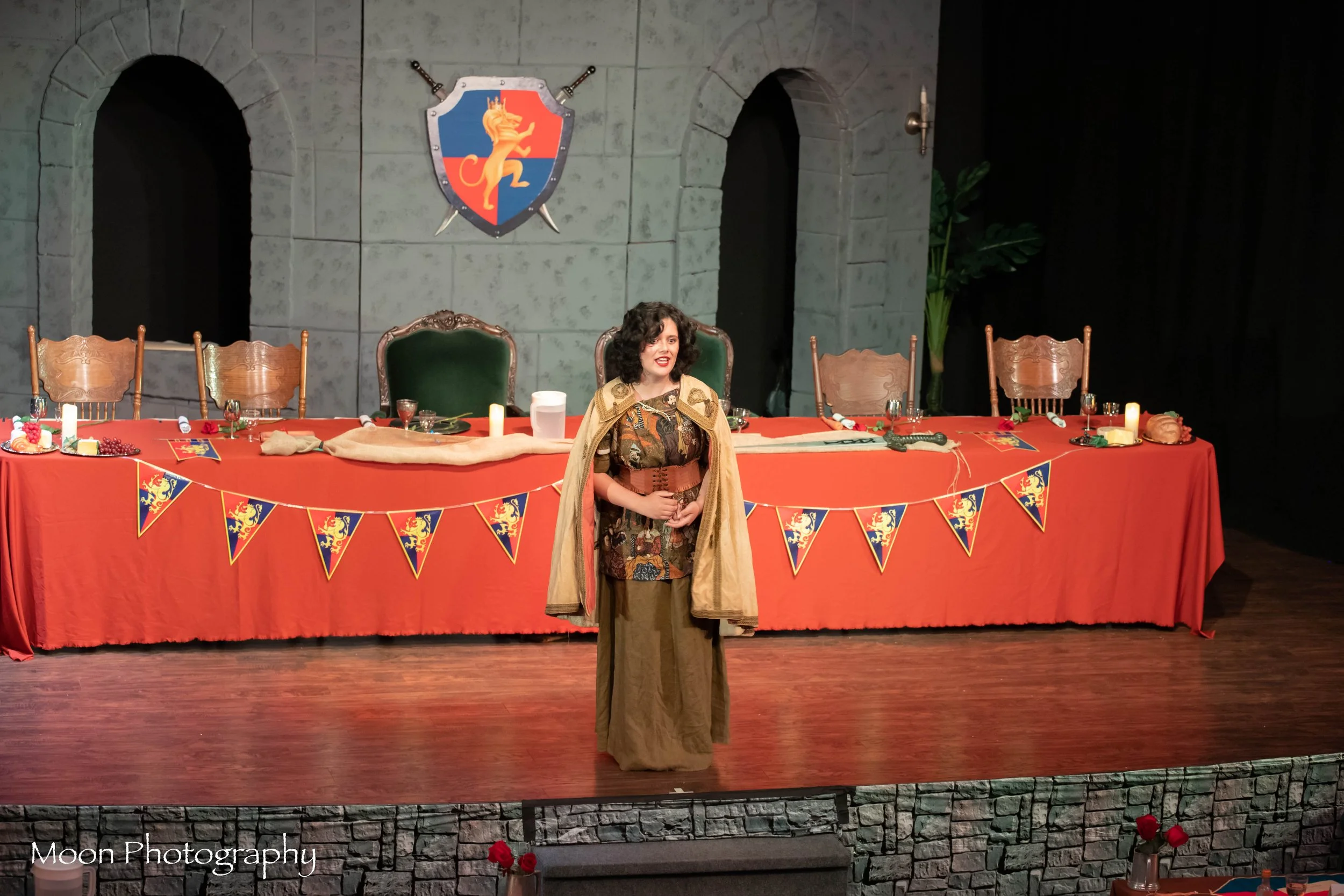 Woman in medieval costume standing on stage with a long red table, green chairs, and medieval banners. The background features gray stone wall and a shield with a lion emblem.
