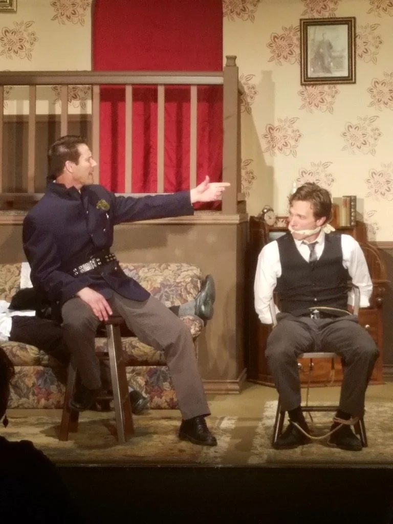 Two men on a stage perform a scene; one man is dressed as a police officer and is sitting on a bench, pointing at the other man who is tied to a chair with rope around his ankles, all in a room with floral wallpaper and a couch.