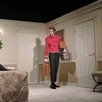 A person standing in a living room holding a box, with a cocktail on a small table nearby.