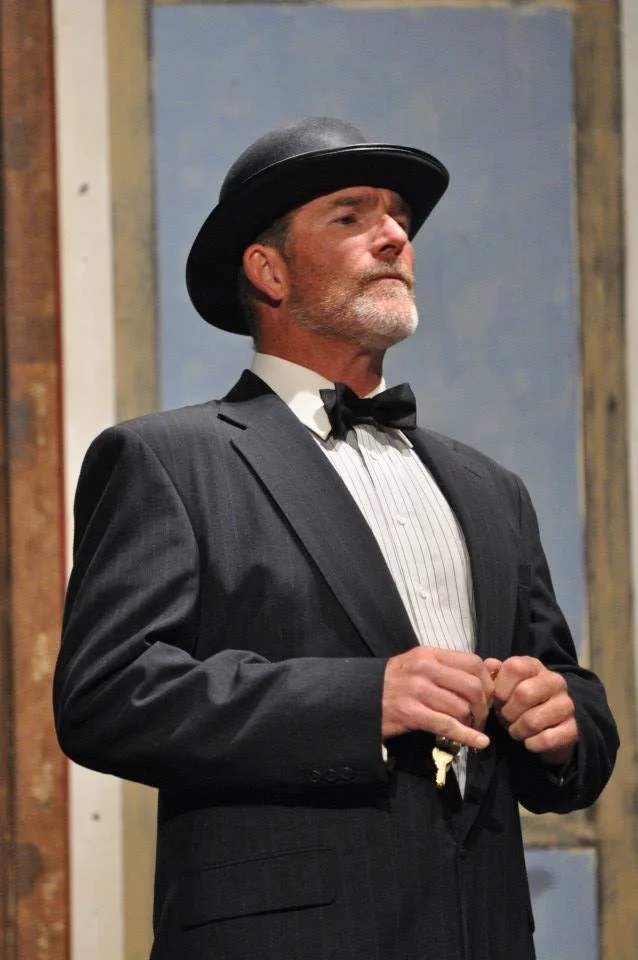 A man dressed in a vintage black tuxedo with a white dress shirt, black bow tie, and a bowler hat, standing with a serious expression against a colorful, rustic backdrop.