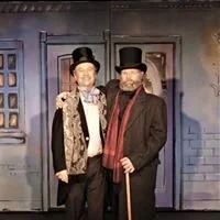 Two men dressed in vintage clothing, wearing top hats and standing in front of a colorful background.