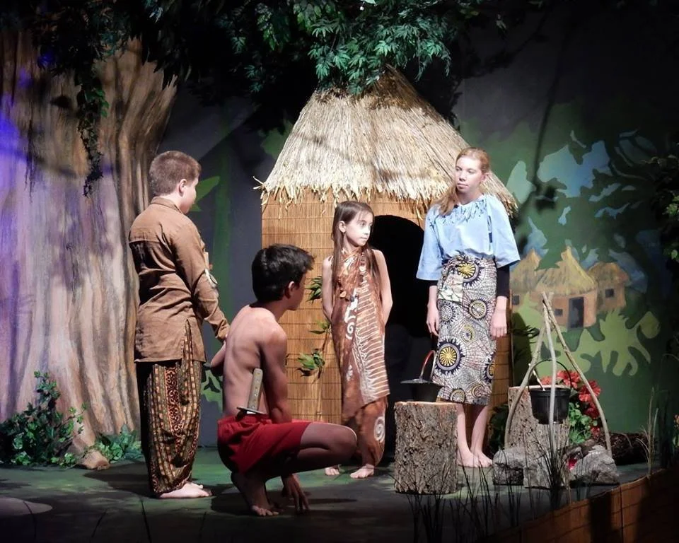 Children performing a play set in a jungle, with a backdrop of trees and a small thatched hut. Four children are dressed in costumes; one girl is in traditional attire, and a girl stands in front of the hut. Two boys are standing and kneeling, one sh