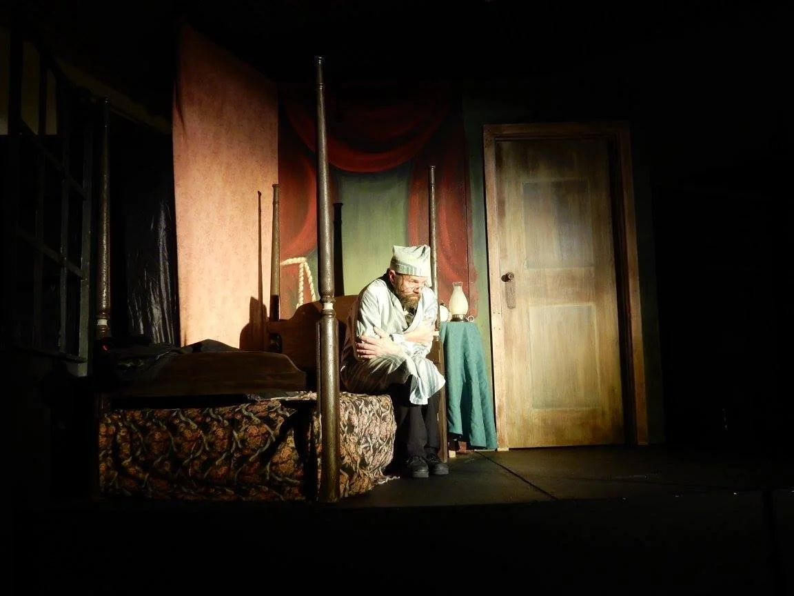 A man dressed in costume sitting on a bed in a theatrical stage setting, with a painted backdrop, a wooden door, and dim lighting.