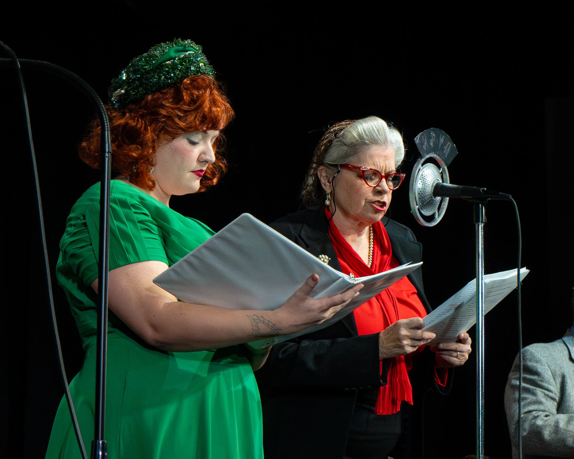 Two women reading from scripts at a microphone, one in a green dress and the other in a black blazer with red blouse and glasses, in a stage setting.