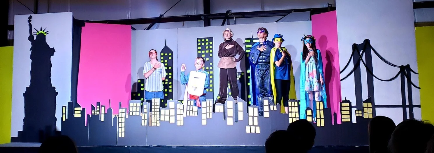 Children performing on a stage with a New York City skyline backdrop, including the Statue of Liberty and a bridge, dressed in costumes representing heroes and everyday characters.