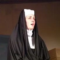 A woman dressed as a nun wearing a black and white habit, appearing to be speaking or singing in front of a wooden background.