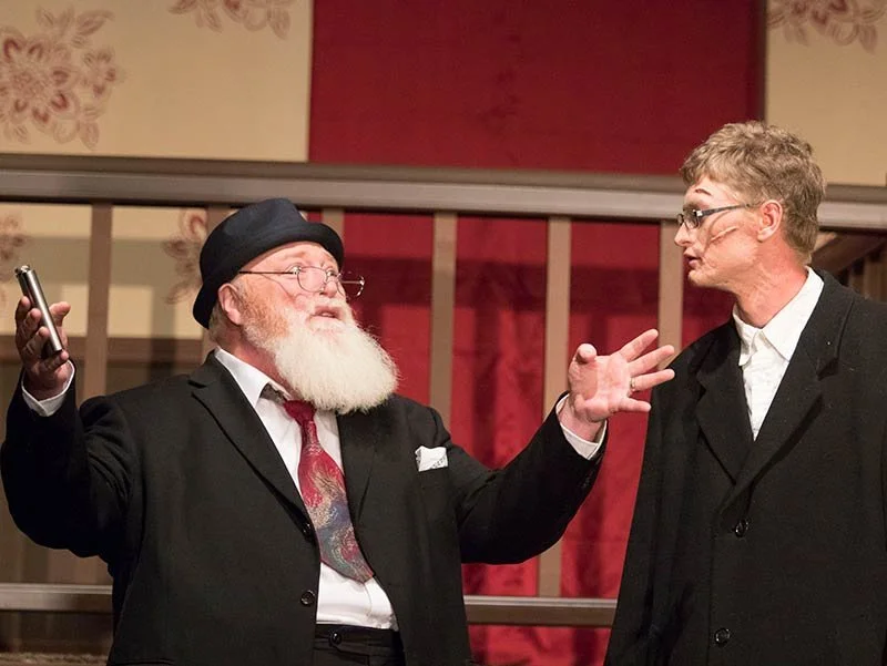 Two men in suits and glasses engaged in conversation, one with a white beard and in a black beret holding a phone, the other with short hair, standing in front of a red and beige patterned background.