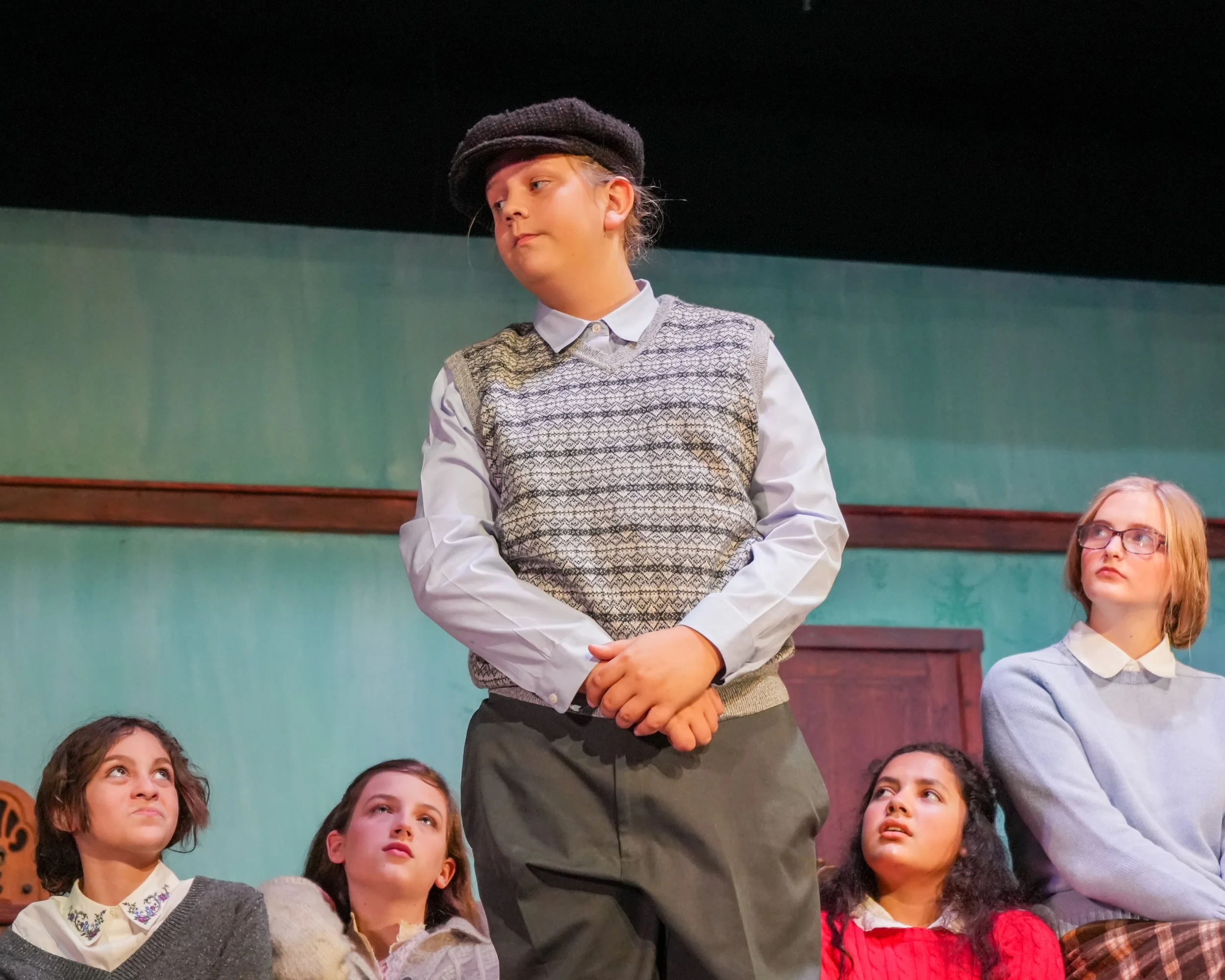 Group of children on stage performing in a school play, with a girl in the center wearing a vintage hat and sweater vest, standing with hands clasped, while the other children sit on the floor looking up at her.