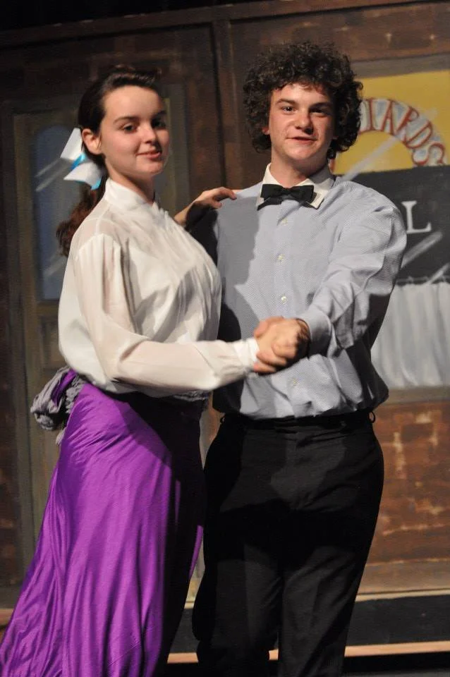 A young woman and a young man dancing together, holding hands in a dance pose, in front of a wooden background. The woman is wearing a white blouse and a purple skirt, with a blue bow in her hair. The man is wearing a light gray shirt with a black bo