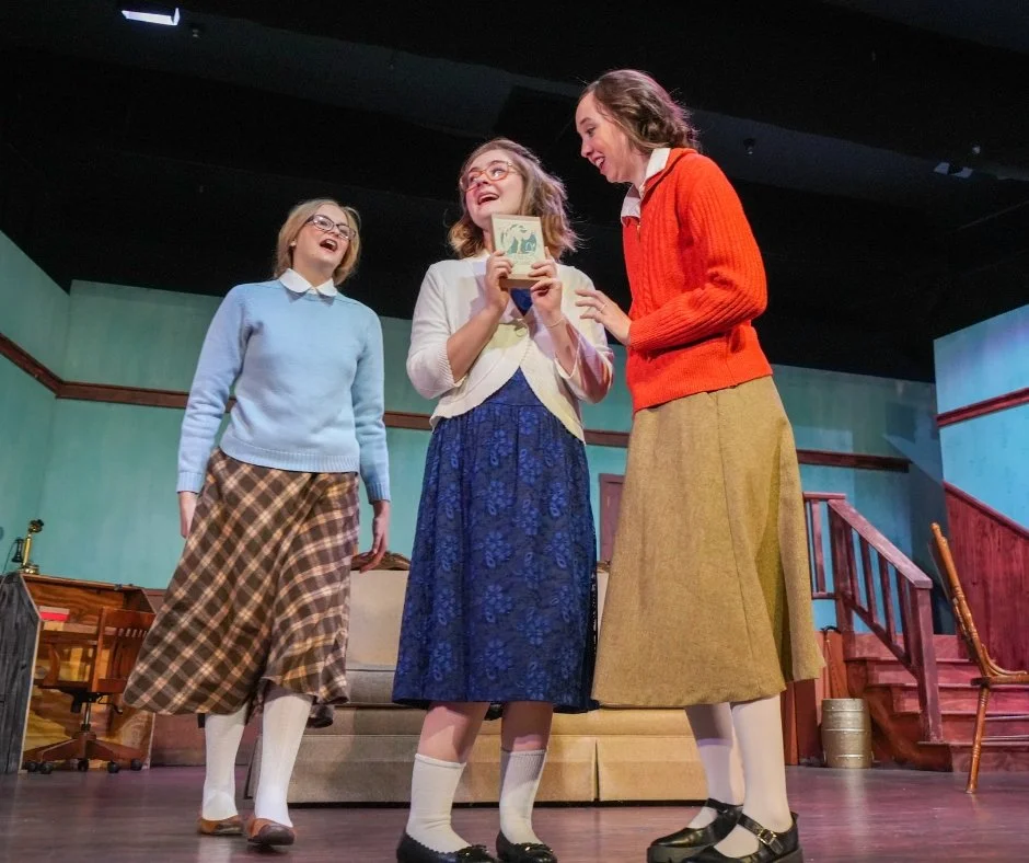 Three women appear to be acting on a stage, holding a book or tablet, with expressions of joy. They are dressed in vintage or retro-style clothing, including long skirts, sweaters, and shoes, suggesting a theatrical performance or play set in an earl