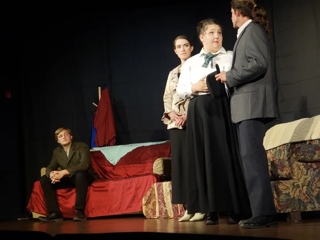A theatrical scene featuring four young actors on stage. One actor sits on a couch, while three stand and appear to be engaged in a conversation. The set includes a red couch, a floral armchair, and a coat hanging on the wall.
