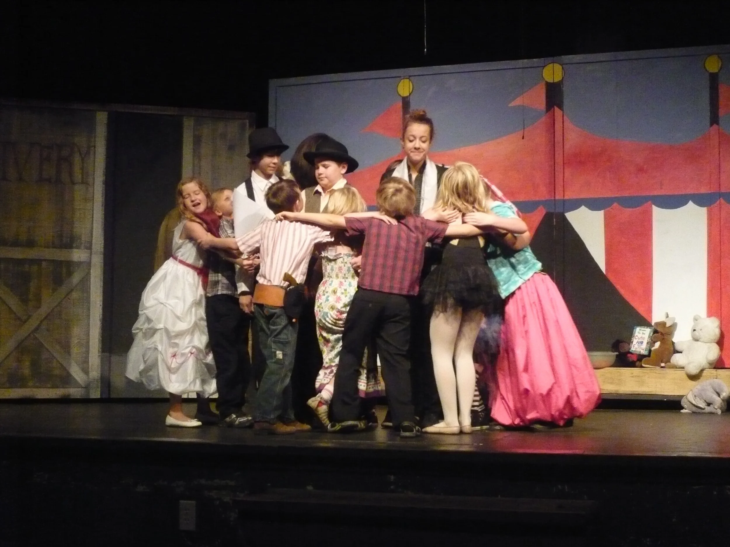 Children dressed in colorful costumes performing on stage at a circus-themed play, embracing in a group hug.