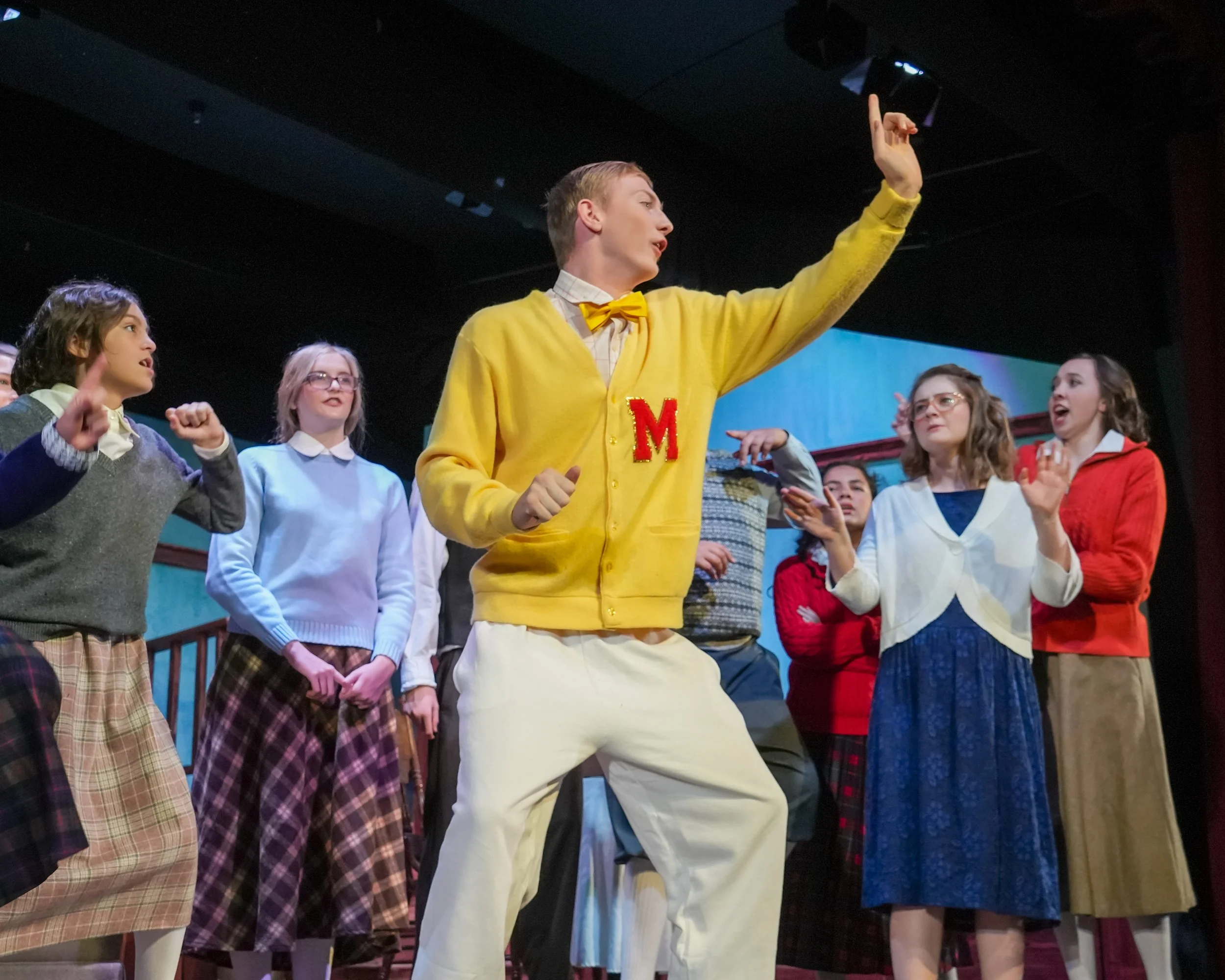 Young actors performing in a school play, dressed in colorful costumes, with a male actor wearing a yellow cardigan with a red letter M, leading the scene.