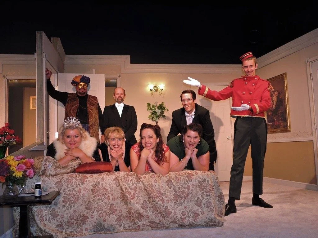 A theatrical play scene with seven actors in colorful costumes posing in a living room set, with some leaning on a floral sofa and others standing behind, smiling and engaging with the audience.