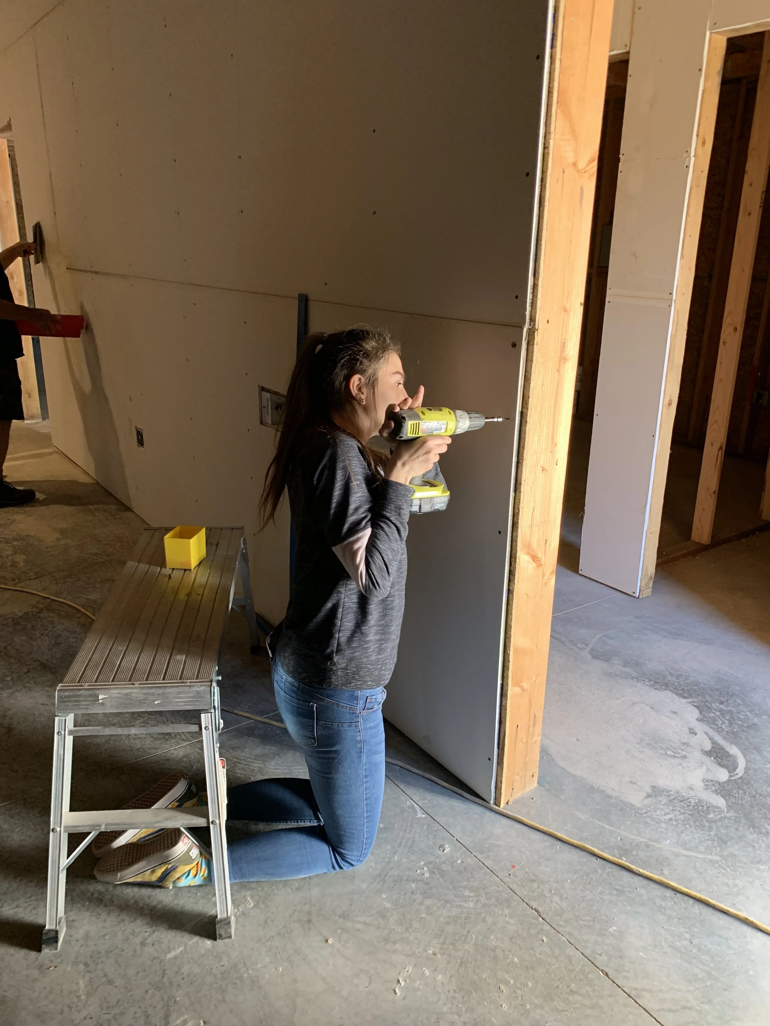 A woman kneeling on the floor using a cordless power drill on a wall under construction in a building.