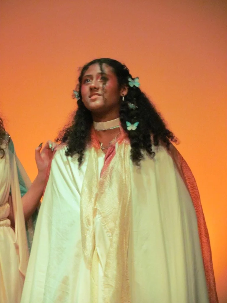 A young woman with dark curly hair decorated with light blue butterfly hair clips, wearing a yellow and gold cape, standing against an orange background, looking emotional.