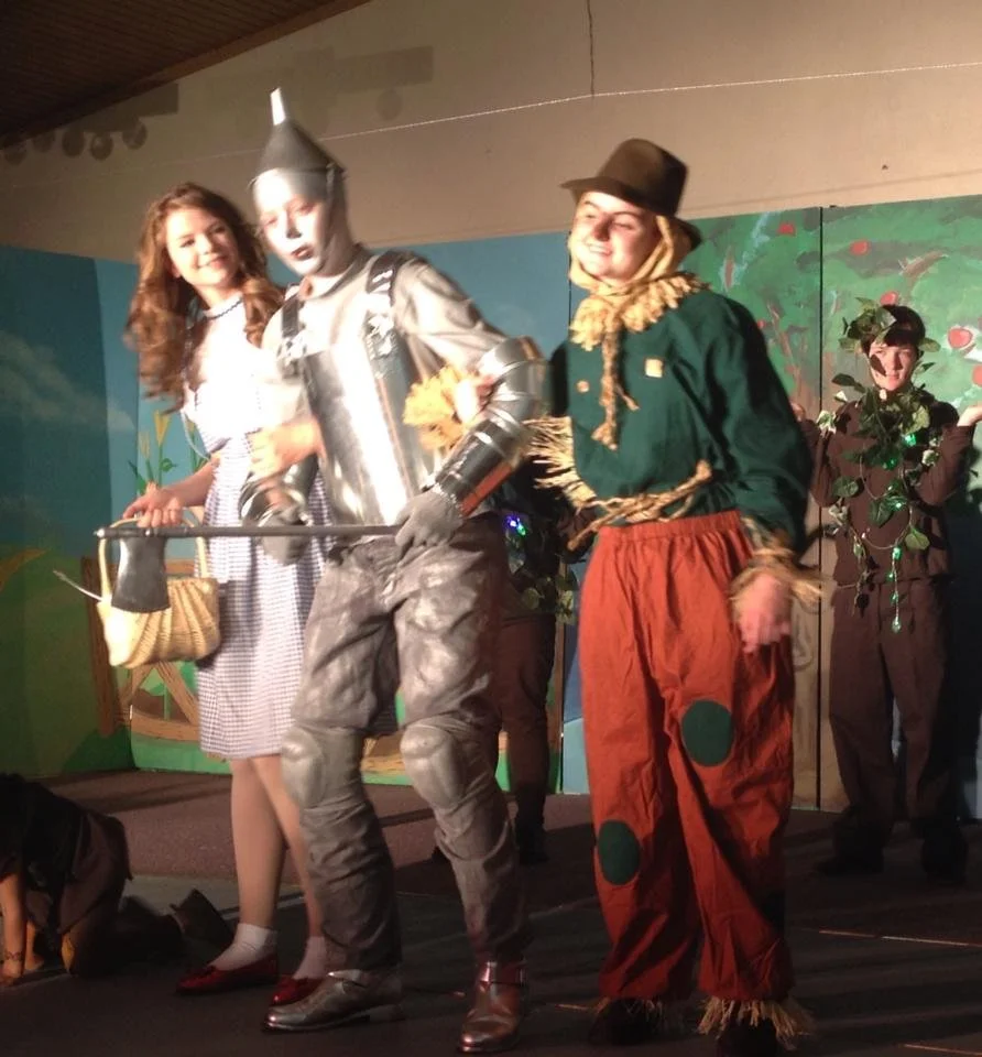 People dressed in costumes perform on stage, including a woman in a gingham dress, a person dressed as the Tin Man from The Wizard of Oz, and a person in a scarecrow costume, with painted backdrop scenery.