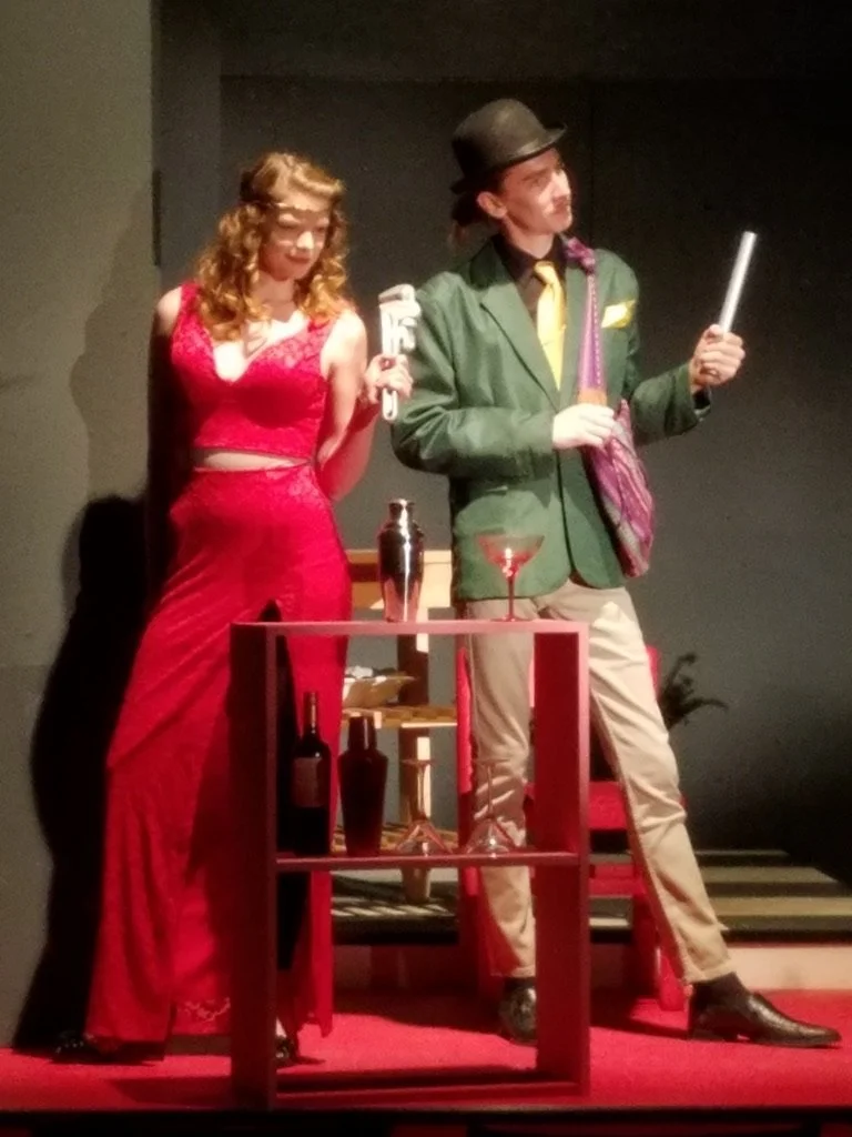 Actors performing a scene on stage with a woman in a red dress and a man in a green blazer and hat, surrounded by bottles and barware.