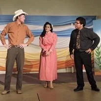 Three people standing on a stage in front of a colorful background, engaged in conversation. The person on the left is wearing a cowboy hat and brown shirt, the person in the middle is wearing a pink dress, and the person on the right is dressed in b
