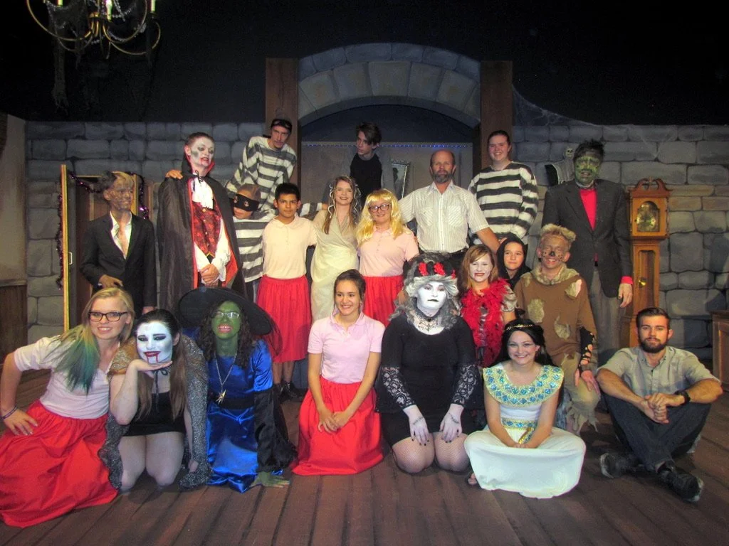 Group of people dressed in theatrical costumes, some as witches, monsters, and characters from fairy tales, on a stage with a spooky castle background.