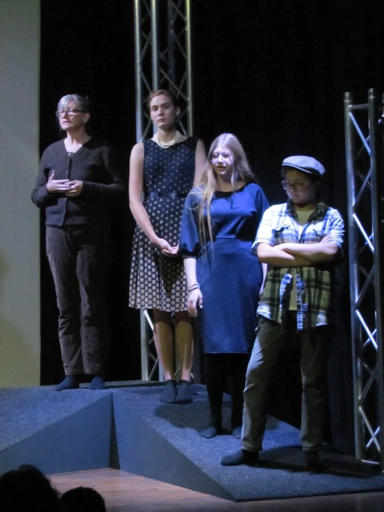 Four young women standing on a stage with a black background and metal trusses, appearing to be part of a theatrical performance.
