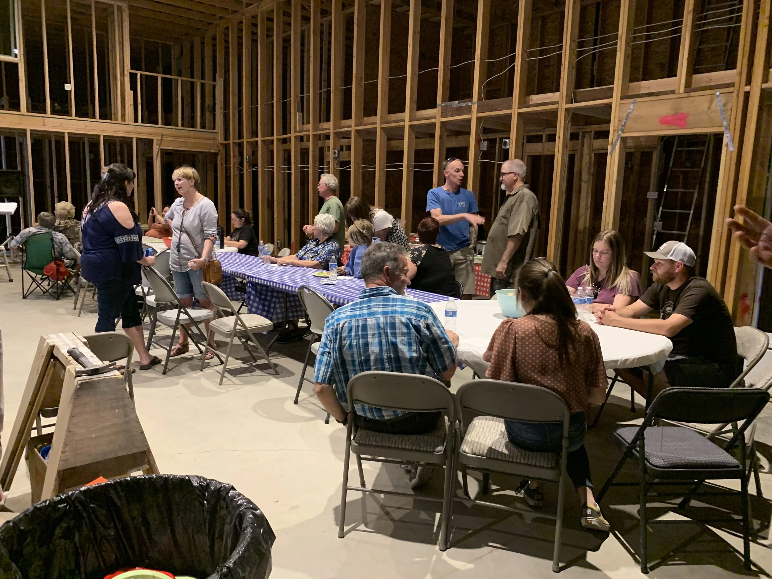 Group of people gathered inside a wooden building, some standing and talking, others sitting at long tables covered with blue and white checkered tablecloths, engaged in conversations.