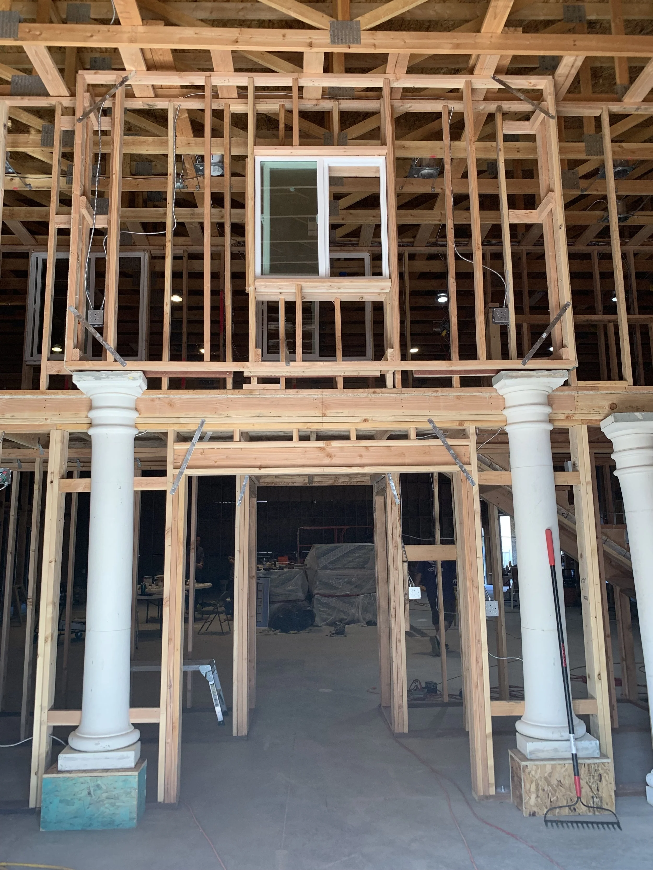 Interior of a building under construction with exposed wooden framing, white decorative columns, and a small upper-level window.