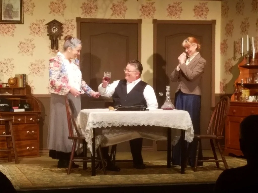 Three people perform in a vintage-style play: a woman standing on the left, a man sitting at a table raising a glass, and a woman standing on the right, smiling, in a room with floral wallpaper and antique furniture.