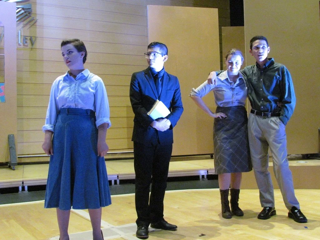 Four young people standing on a stage, posing for a photo, dressed in semi-formal attire, with a wooden background and a partial word 'LEY' visible on the wall.