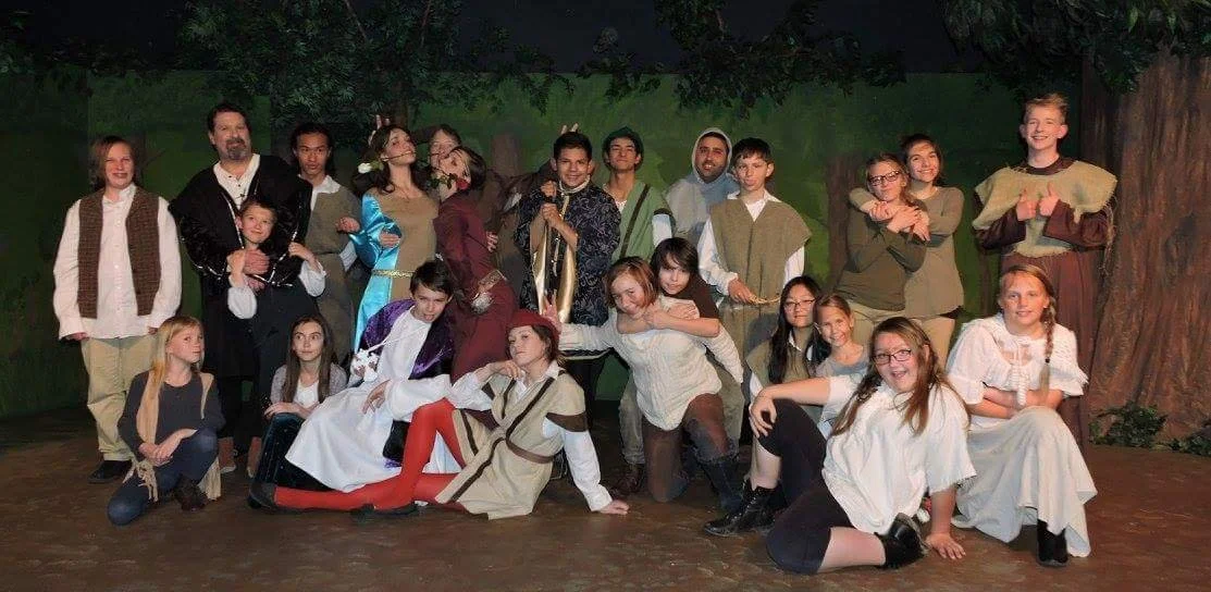 Group of children and adults in costume on a stage, performing a theatrical play, with a forest backdrop.