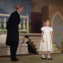 A woman and a young girl standing on a stage set resembling a living room with a staircase. The woman is dressed in dark clothing, and the girl is wearing a white dress with short sleeves.