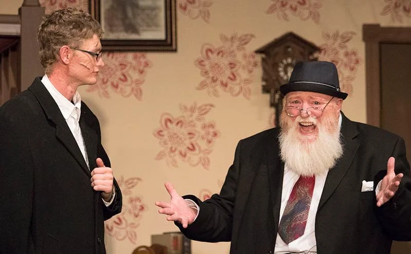 Two men dressed in formal attire, one with glasses and a hat, are having a lively conversation in a room with floral wallpaper.