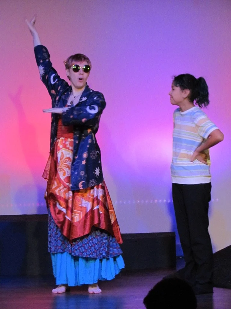 Person dressed in cosmic-themed costume with sunglasses performing on stage while a young girl watches with interest.