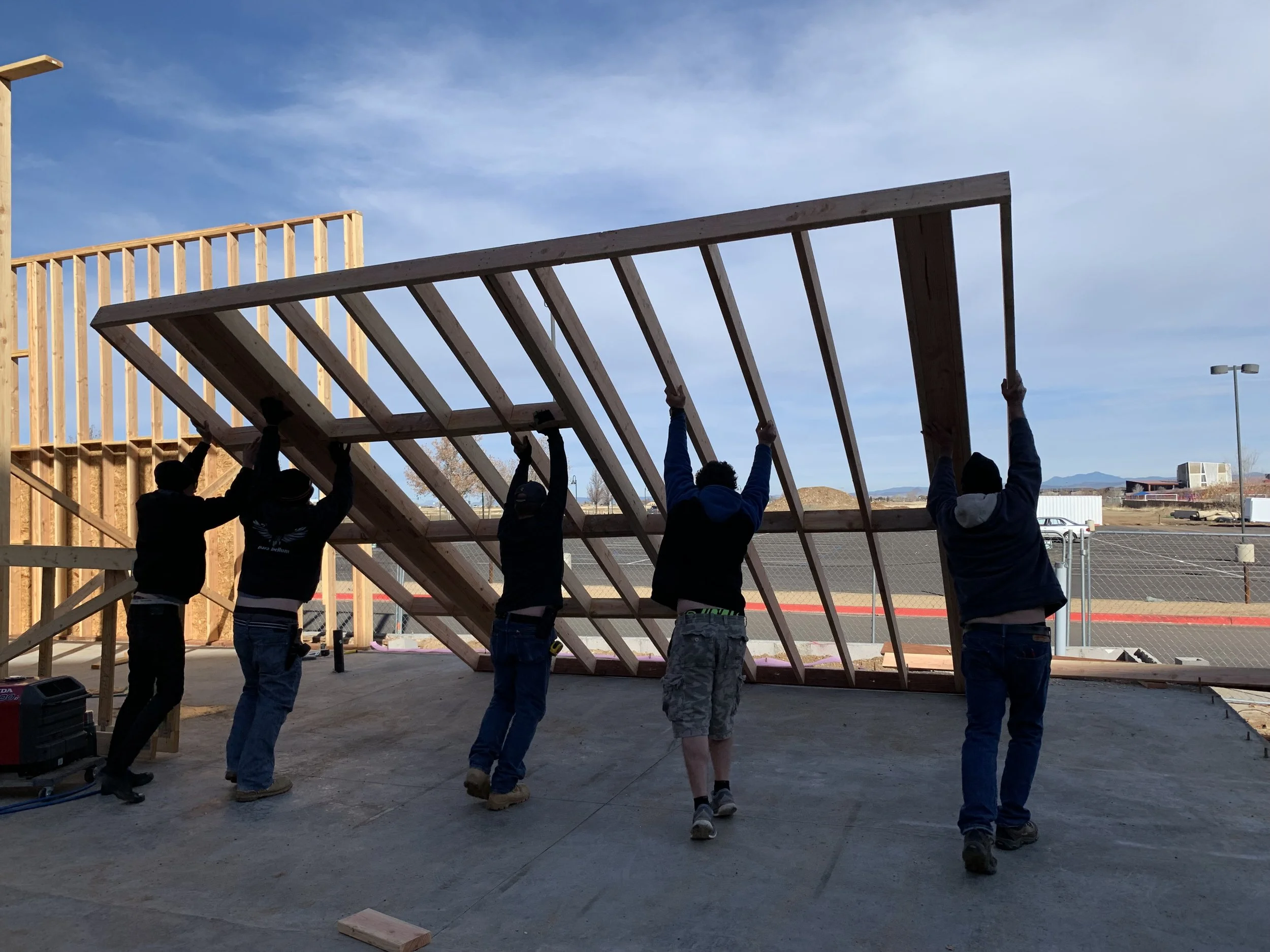 Five workers are lifting and tilting a large wooden roof frame into position during construction on a building site outdoors.