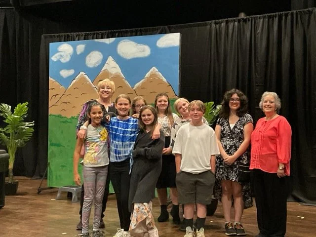 Group of children and two women standing on stage in front of a painted backdrop of mountains, clouds, and trees, with black curtains and potted plants on either side.