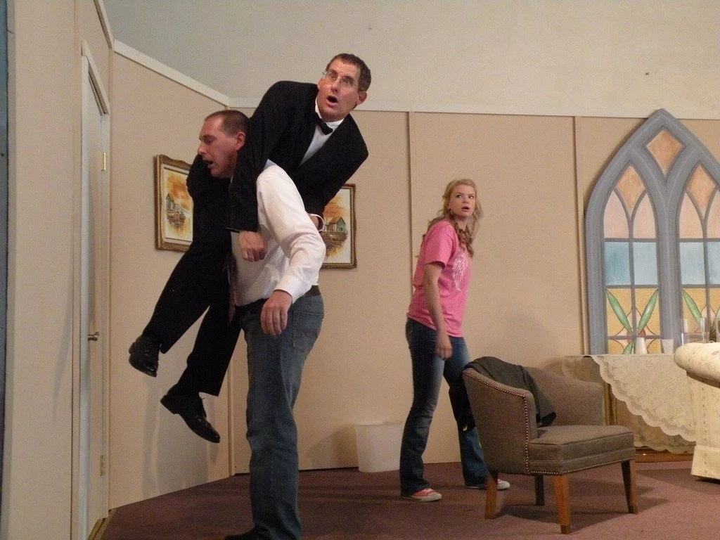 A man in a suit and glasses is carrying another man in a white shirt and black jacket on his back, both with surprised expressions, in a room with a woman in a pink shirt and jeans looking shocked, with furniture and a stained glass window in the bac