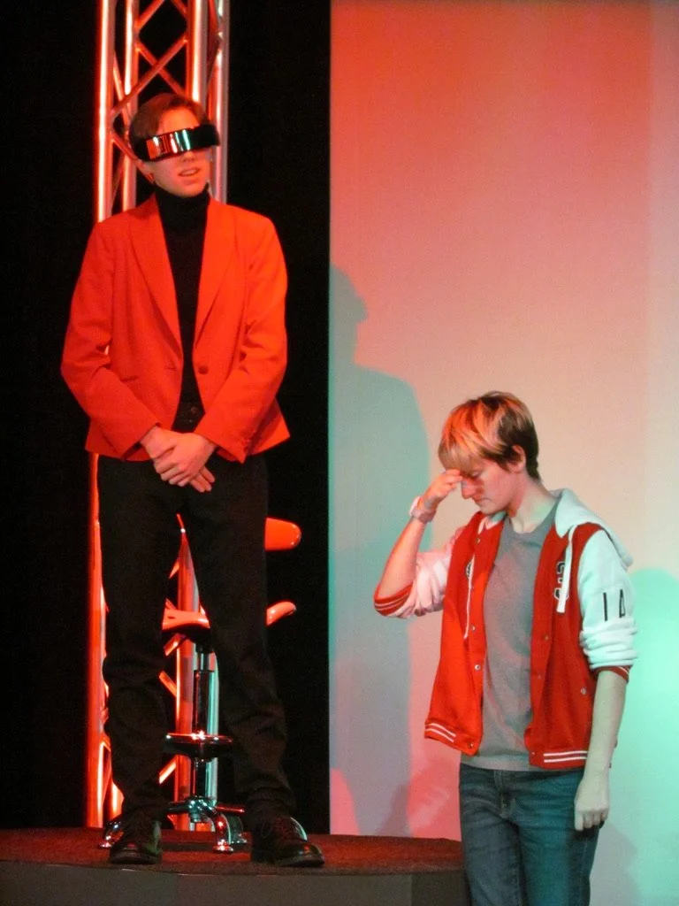 Two individuals on a stage, one standing with a VR headset, the other with a hand on their face, wearing casual clothing and a red jacket.