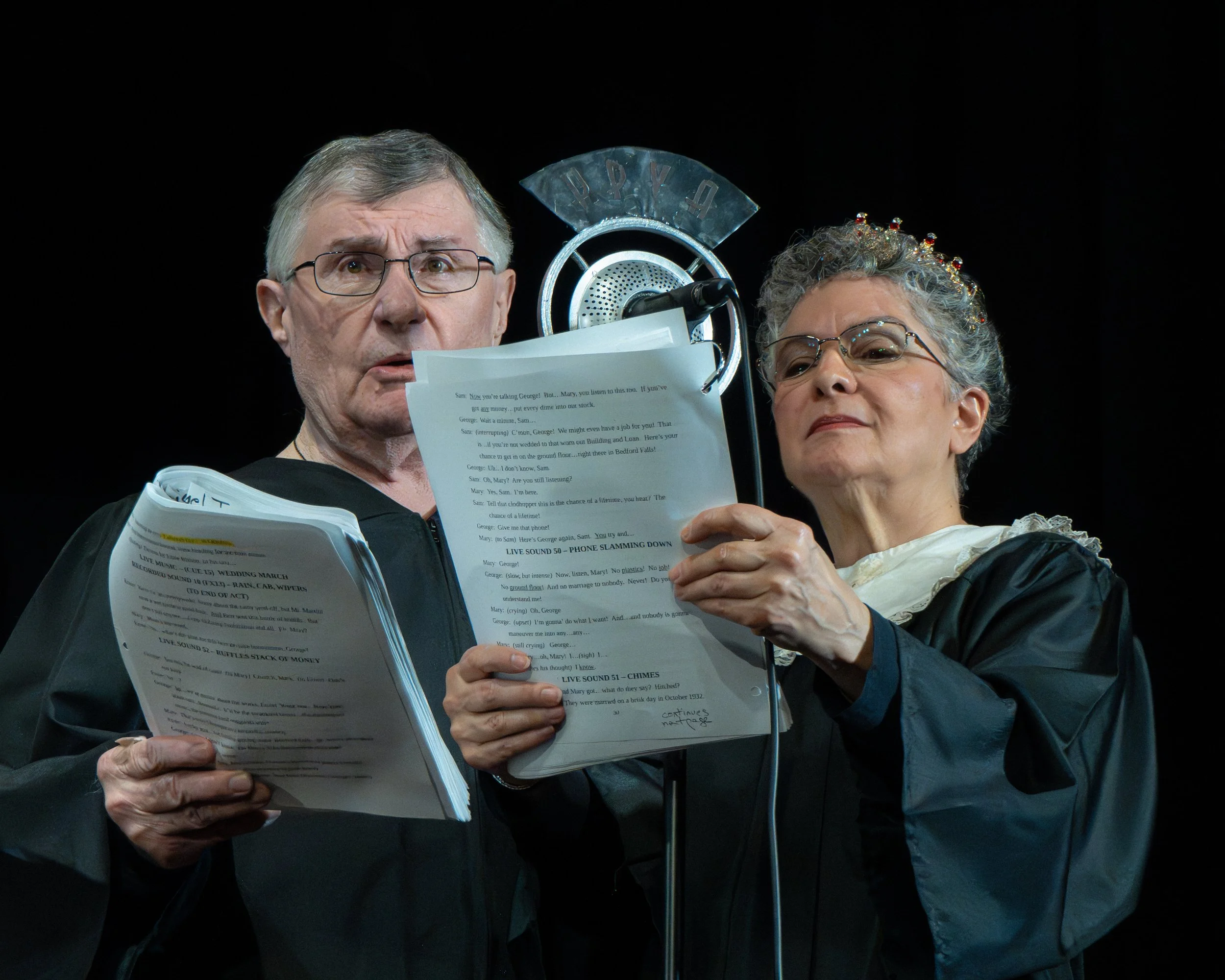 Two older actors dressed as judges stand at a microphone, reading from scripts during a theatrical performance.