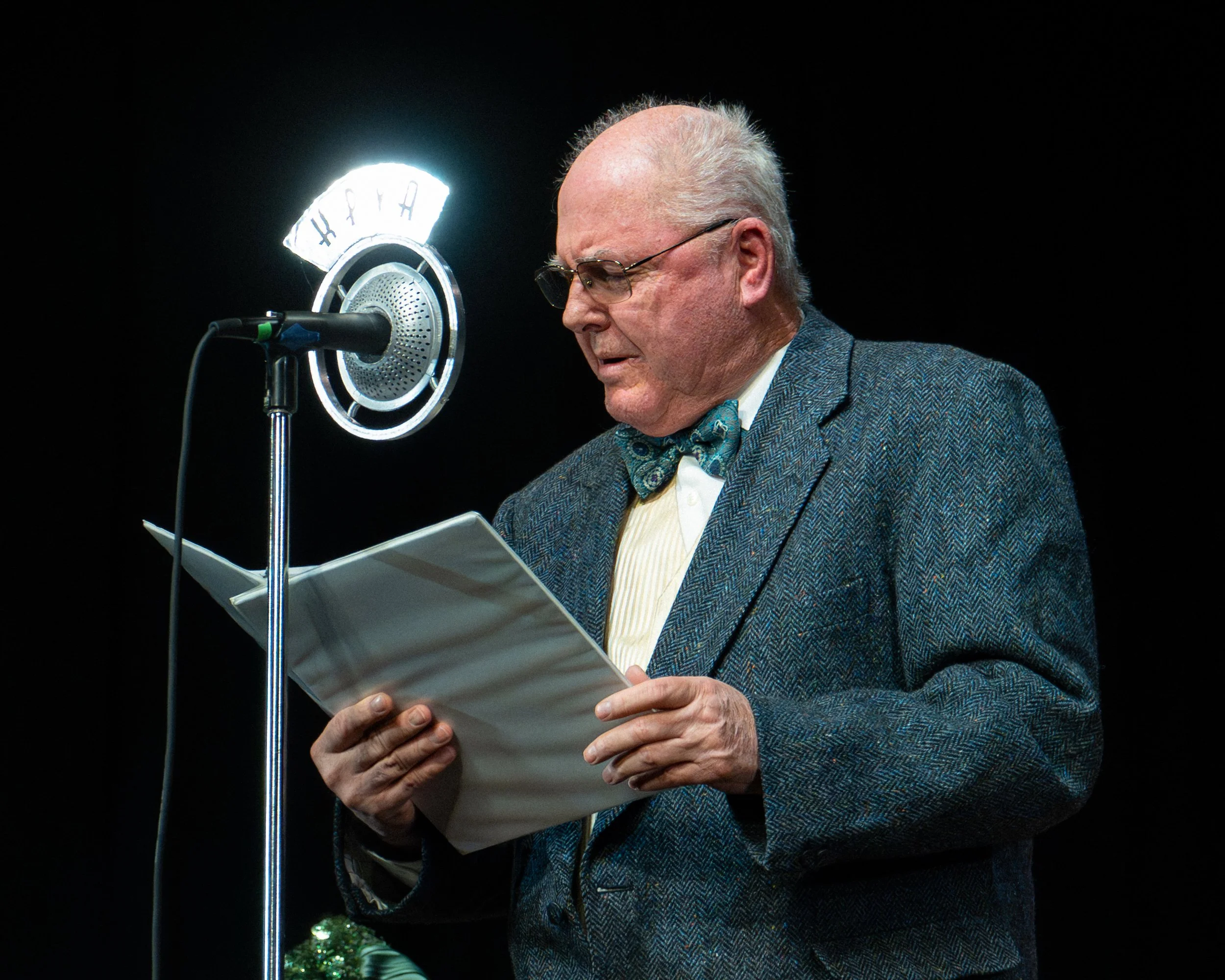 Elderly man in a suit with a bow tie reading from papers at a microphone with a bright light behind it.