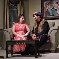 A young woman in a pink dress sitting on a sofa and talking to a man dressed as Aladdin in a blue turban and vest, in a living room setting.