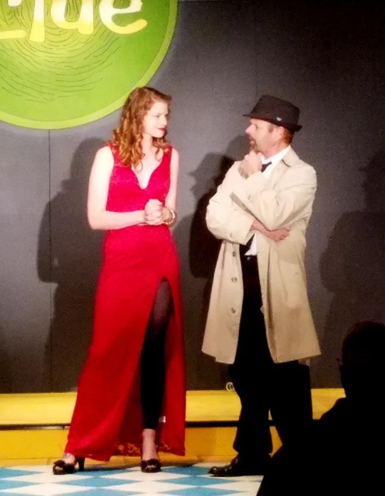 A woman in a red dress and a man in a beige trench coat and black hat standing on a stage, engaged in a conversation. The woman has curly red hair and is wearing black tights and heels, while the man has a beard and is holding his chin with his hand.