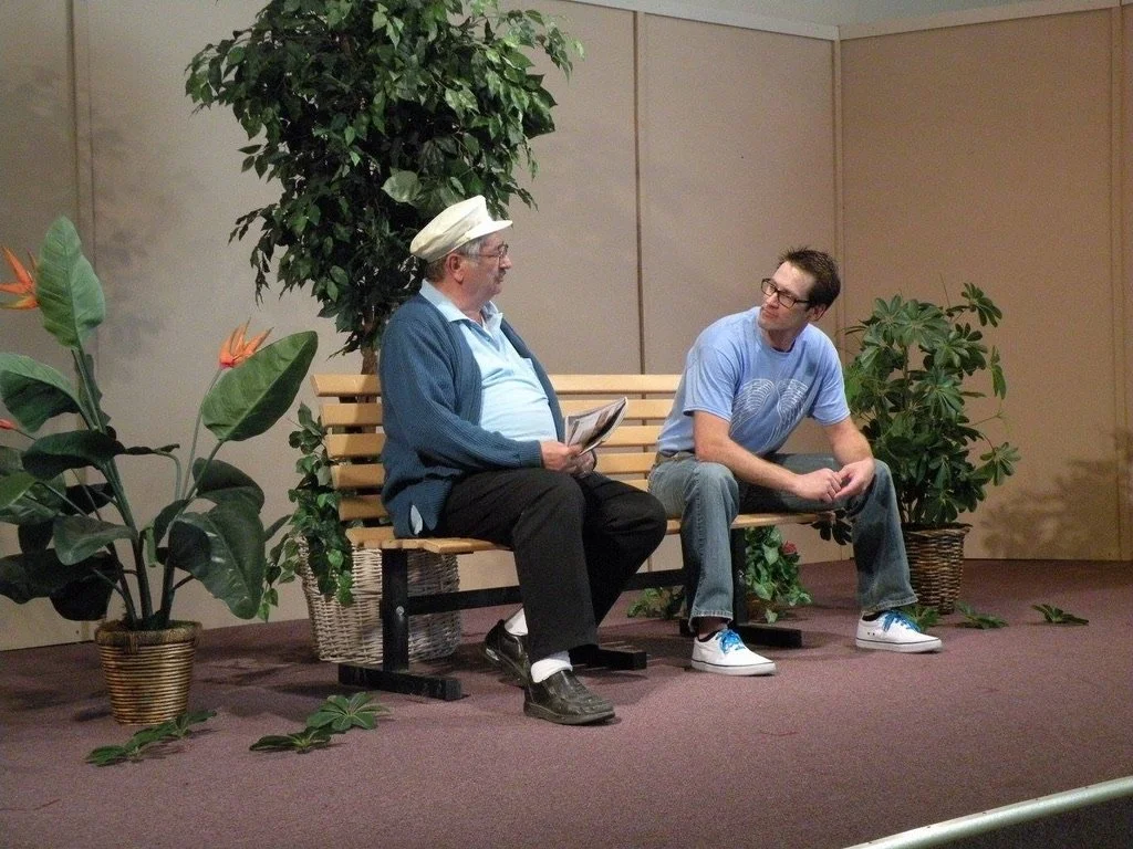 Two men sitting on a wooden bench, engaged in conversation, surrounded by potted plants indoors.