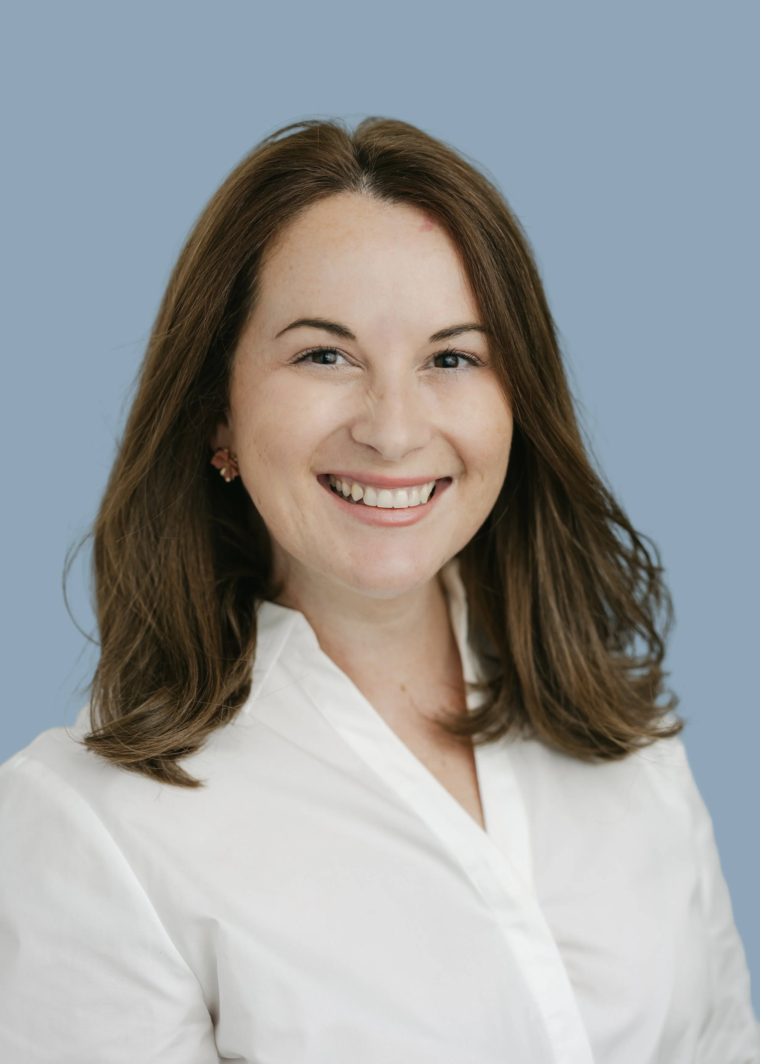 A woman with light skin, shoulder-length brown hair, and brown eyes smiling, wearing a white shirt and floral earrings, against a blue background.